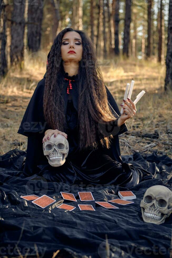 bruja en negro vestir con capa y capucha. posando en pino bosque. sentado en oscuro cobija con adivinación tarjetas y calaveras en él. lleno longitud. foto