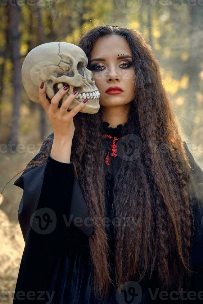 Witch in black, long dress with cape and hood. Posing in smoke pine forest. Holding a skull. Spells, magic and witchcraft. Close-up. photo
