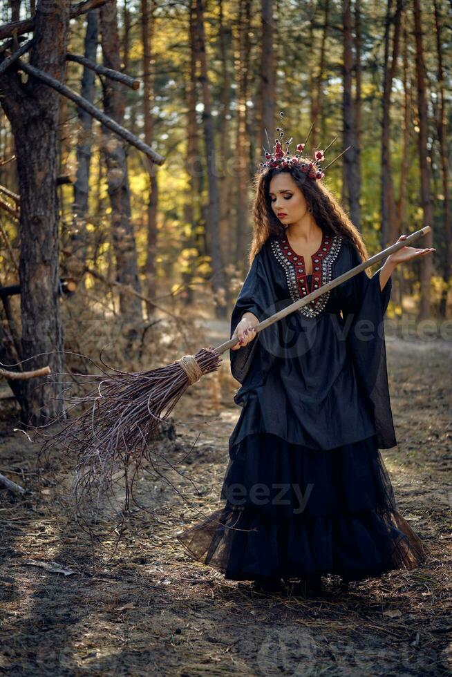 Witch in black, long dress, with red crown in her long hair. Posing with broom in pine forest. Spells, magic and witchcraft. Full length portrait. photo