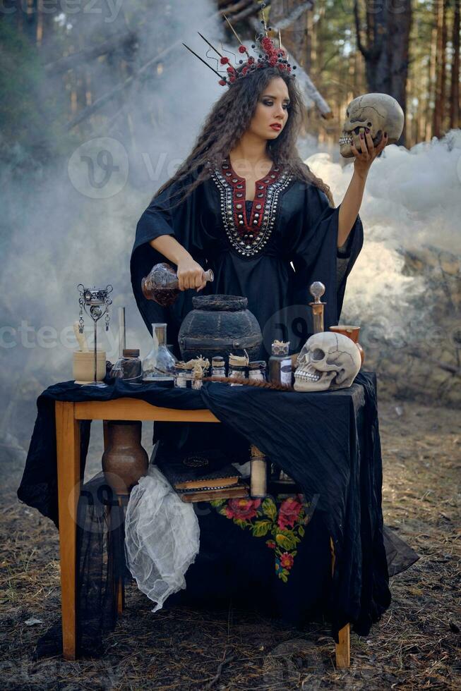 Witch in black, long dress, with red crown in hair. Posing in pine forest, holding skull, making potion. Spells and witchcraft. Full length, smoke. photo