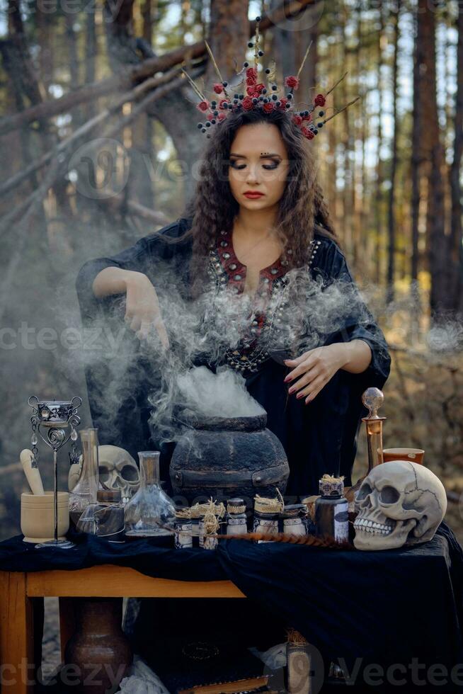 Witch in black, long dress, with red crown in her long hair. Posing in pine forest. Making a magic potion. Spells and witchcraft. Close-up, smoke. photo