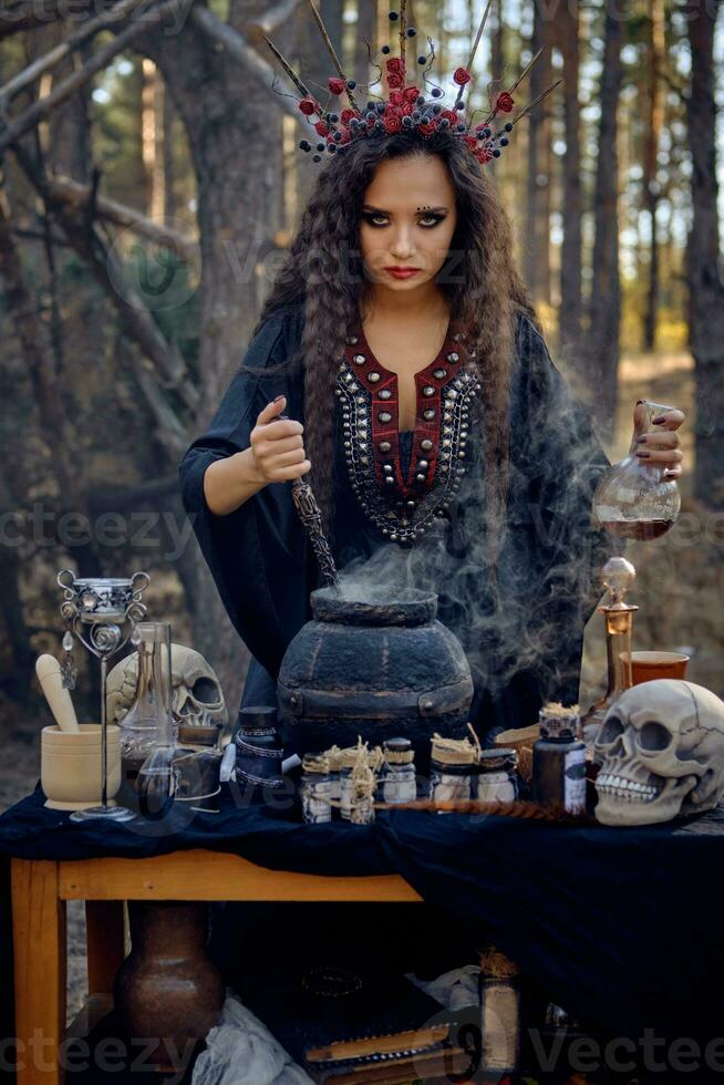 Witch in black, long dress, with red crown in her long hair. Posing in pine forest. Making a magic potion. Spells and witchcraft. Close-up, smoke. photo