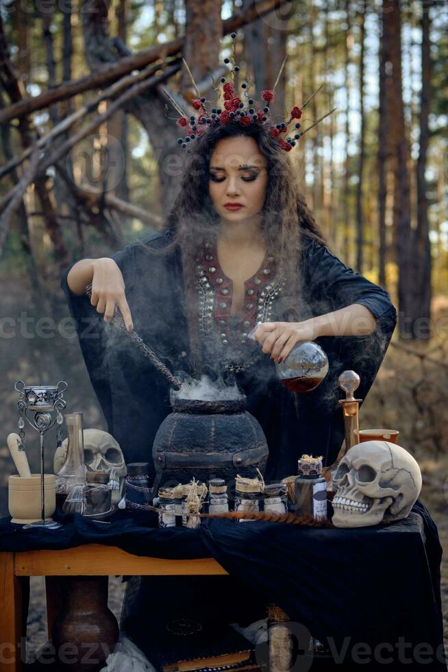 Witch in black, long dress, with red crown in her long hair. Posing in pine forest. Making a magic potion. Spells and witchcraft. Close-up, smoke. photo