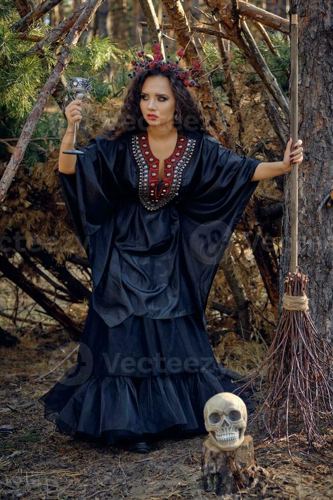 Witch in black, long dress, with red crown in her long hair. Posing with broom and skull in pine forest. Spells, magic and witchcraft. Full length. photo