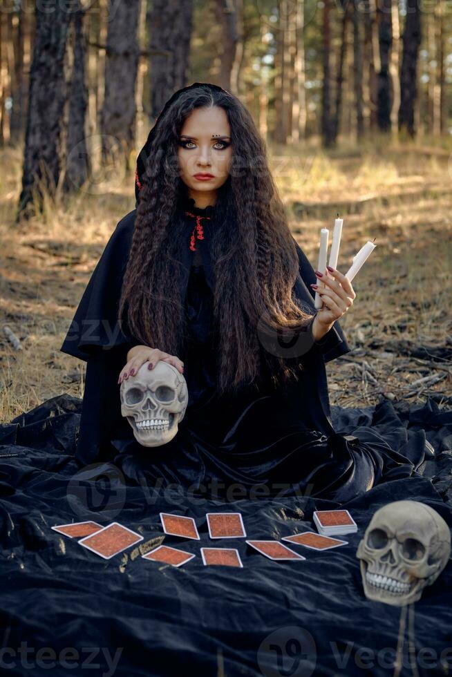 Witch in black dress with cape and hood. Posing in pine forest. Sitting on dark blanket whith fortune-telling cards and skulls on it. Full length. photo