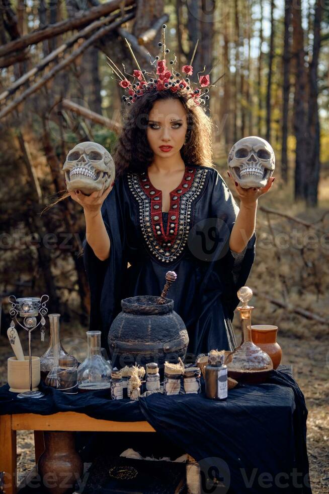 Witch in black, long dress, with red crown in her long, curly hair. Posing in pine forest. Holding skulls. Spells, magic and witchcraft. Close-up. photo