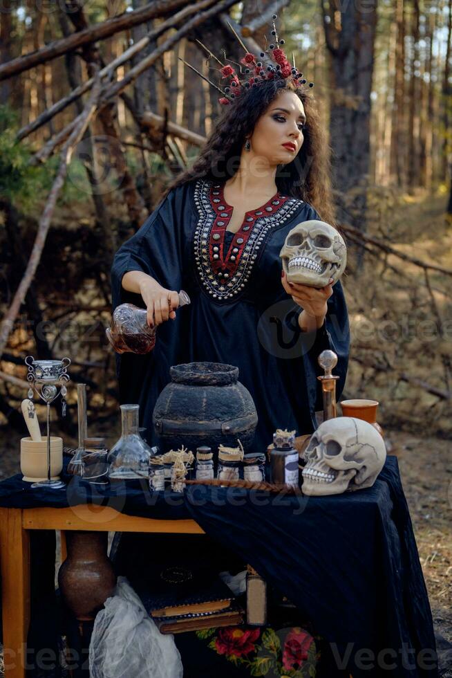 Witch in black, long dress, with red crown in her hair. Posing in pine forest, holding skull, making potion. Spells and witchcraft. Close-up. photo