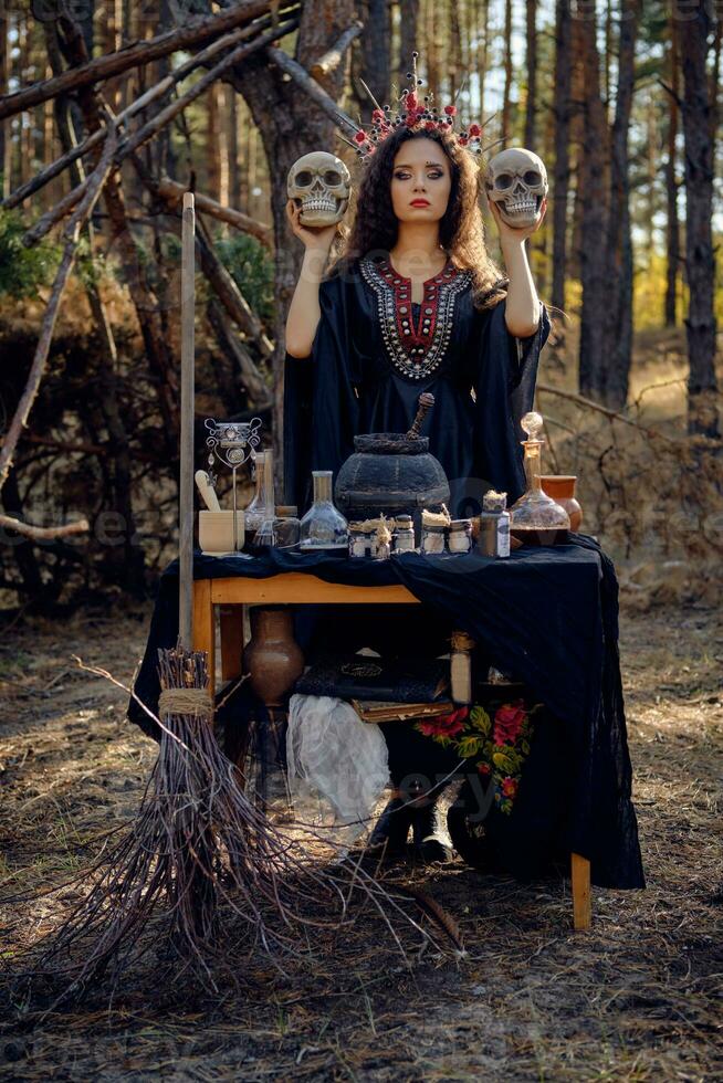 Witch in black, long dress, with red crown in her long hair. Posing in pine forest. Holding skulls. Spells, magic and witchcraft. Full length. photo