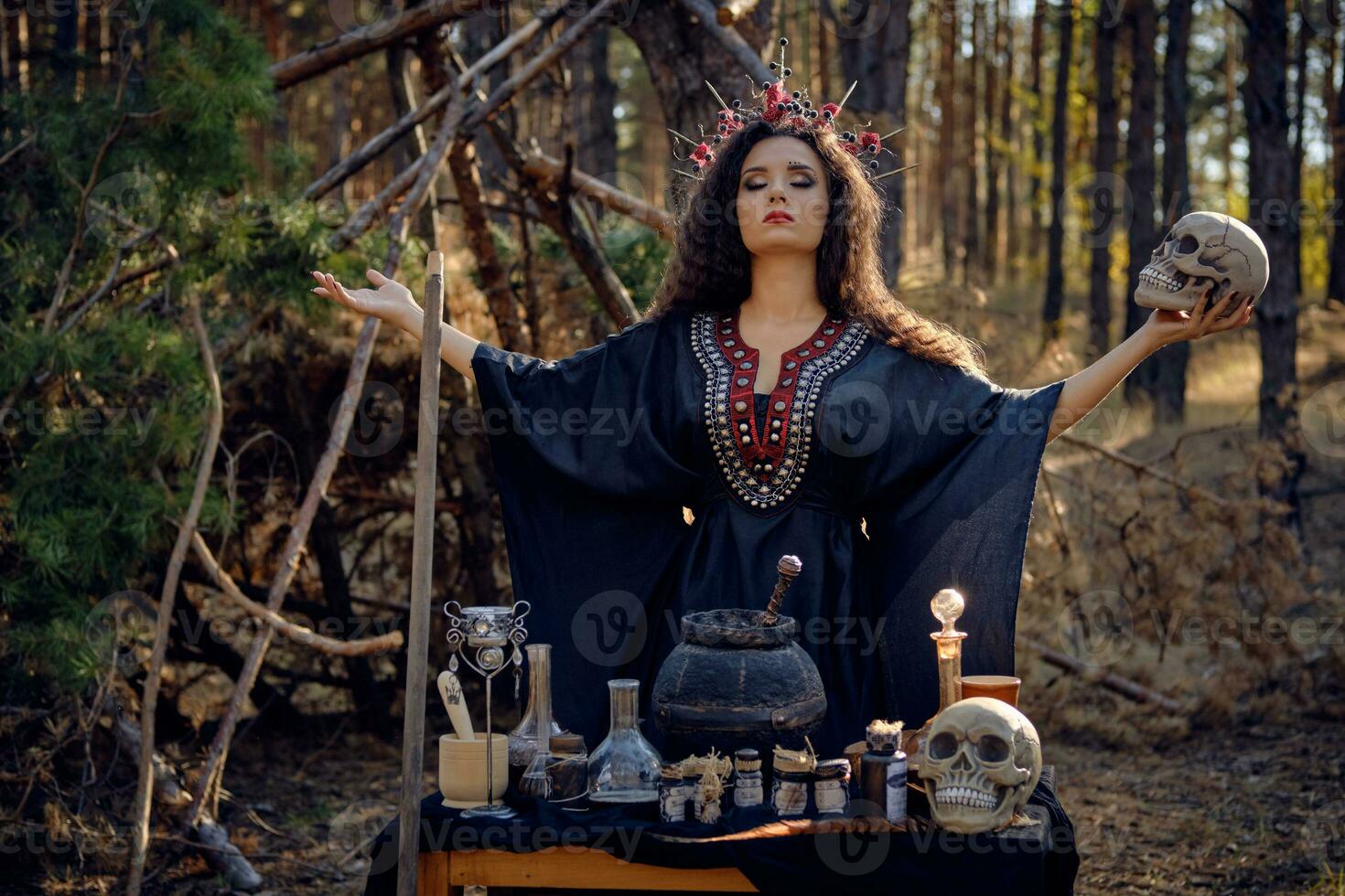 Witch in black, long dress, with red crown in her long, curly hair. Posing in pine forest. Holding skull. Spells, magic and witchcraft. Close-up. photo