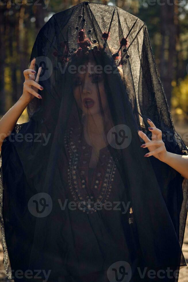 Witch in black, long dress, with red crown in her long, curly hair under a black veil. Posing in pine forest. Spells, magic and witchcraft. Close-up. photo