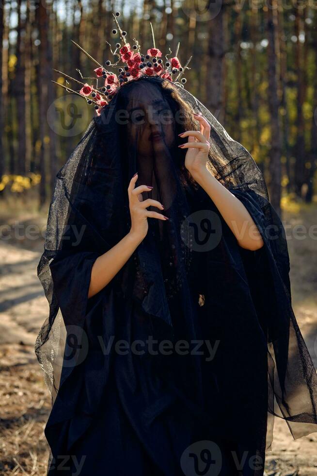 Witch in black, long dress, with red crown in her long, curly hair under a black veil. Posing in pine forest. Spells, magic and witchcraft. Close-up. photo