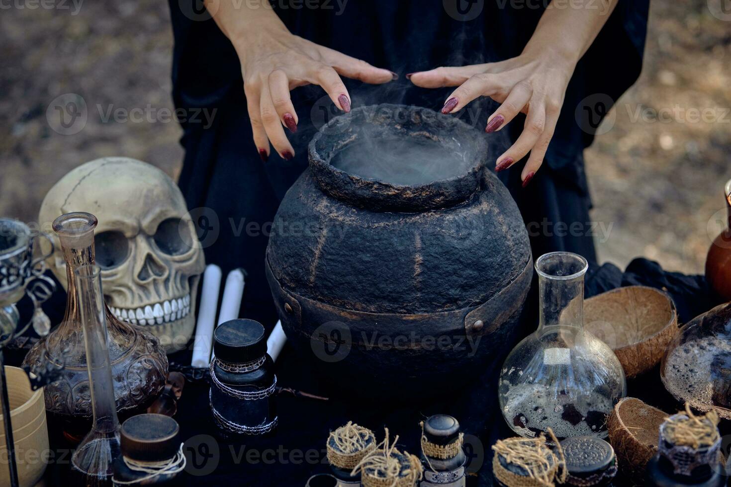Witch in black dress is making a magic potion in a dark pot. Posing in pine forest. Spells and witchcraft. Close-up, smoke. photo