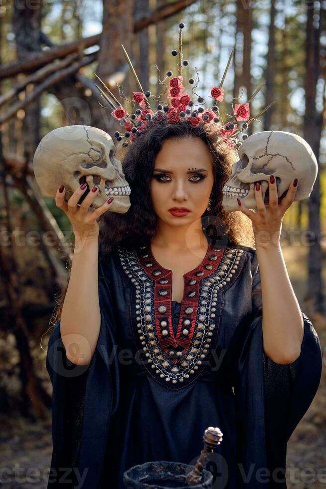 Witch in black, long dress, with red crown in her long, curly hair. Posing in pine forest. Holding skulls. Spells, magic and witchcraft. Close-up. photo