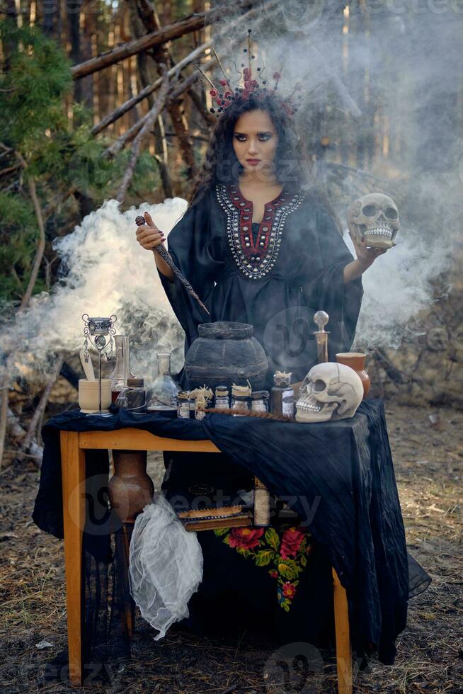 Witch in black, long dress, with red crown in hair. Posing in pine forest, holding skull, making potion. Spells and witchcraft. Full length, smoke. photo