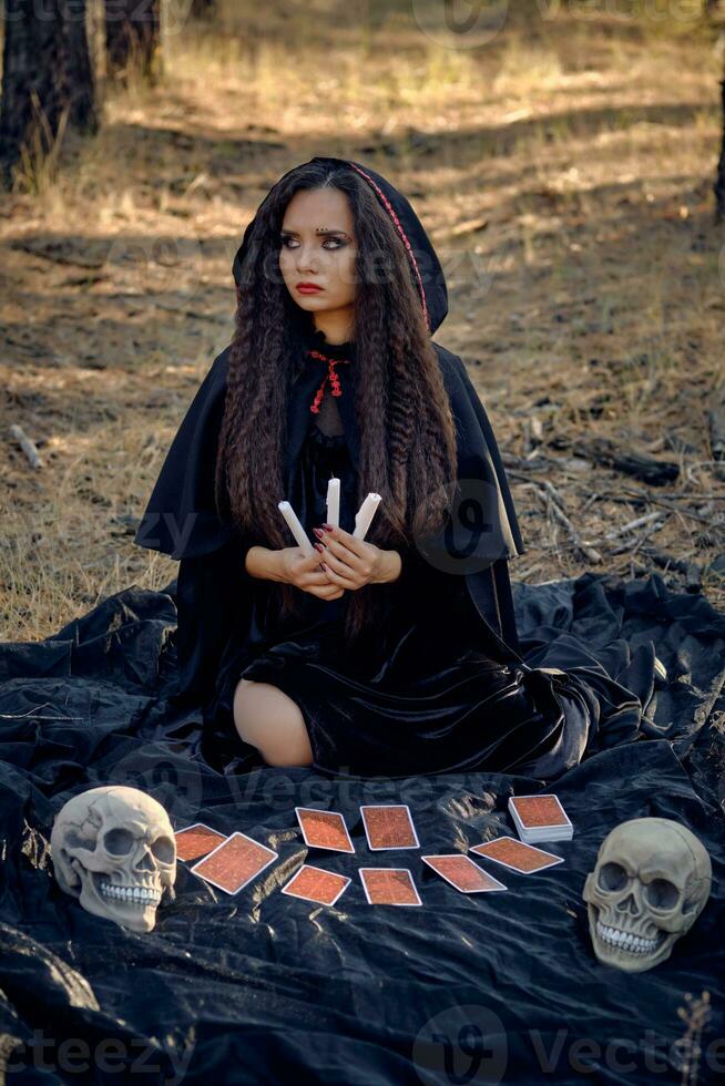 Witch in black dress with cape and hood. Posing in pine forest. Sitting on dark blanket whith fortune-telling cards and skulls on it. Full length. photo