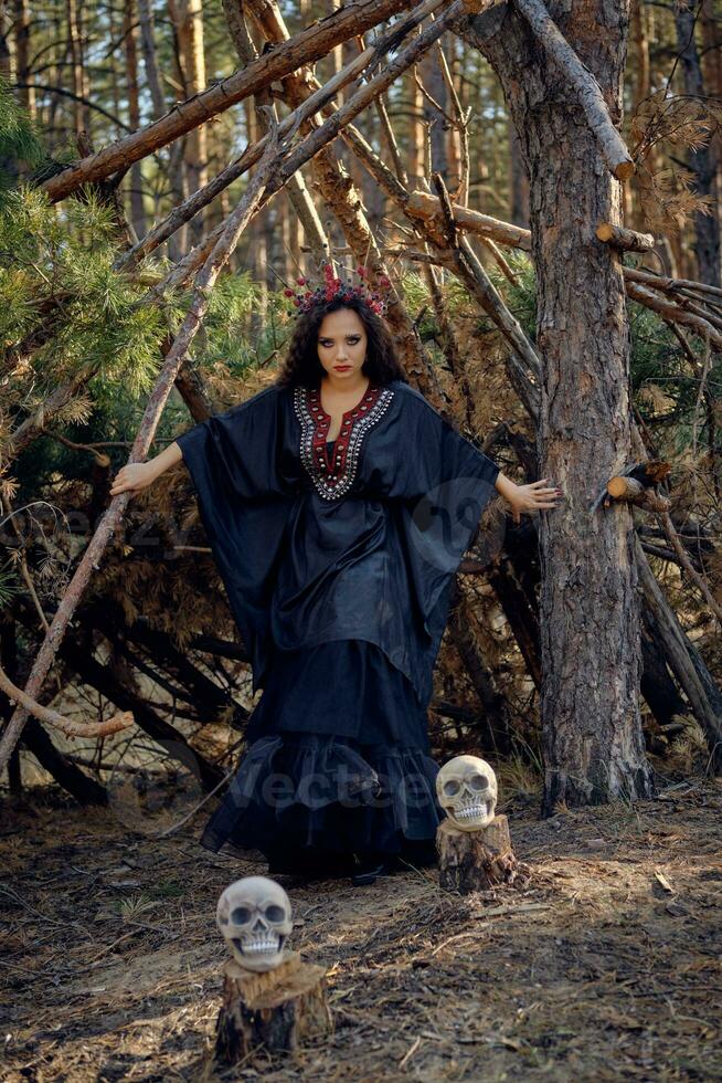 bruja en negro, largo vestido, con rojo corona en su largo cabello. posando con Escoba y cráneo en pino bosque. hechizos, magia y brujería. lleno longitud. foto