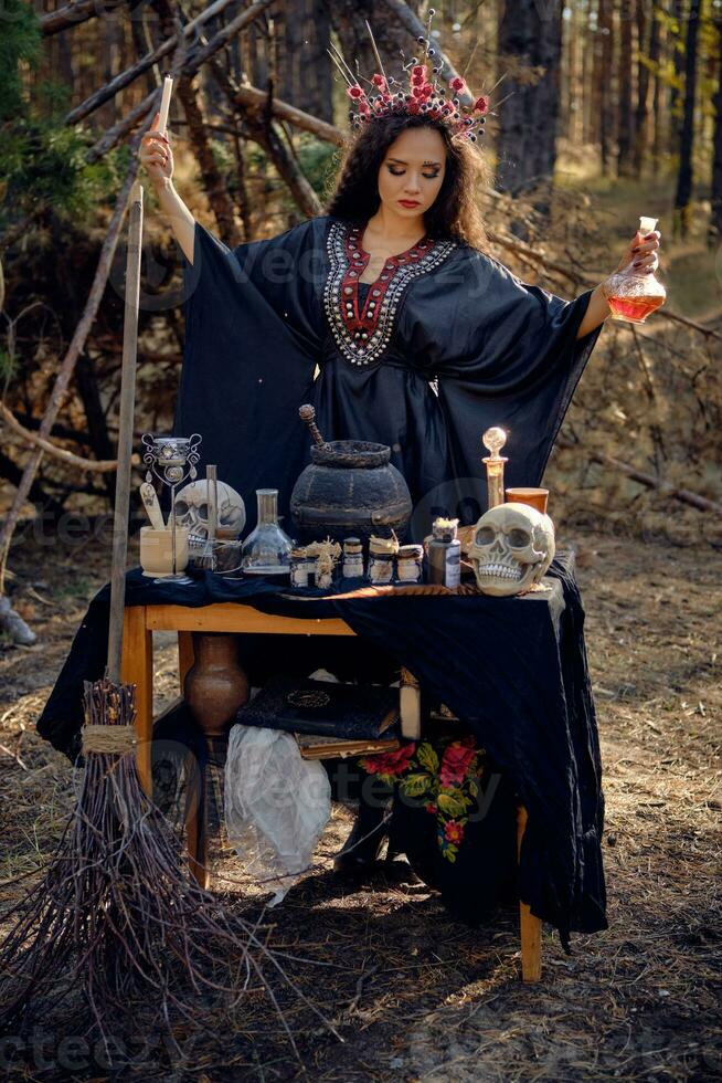 hermosa bruja en negro, largo vestido, con rojo corona en su largo cabello. posando en pino bosque. hechizos, magia y brujería. lleno longitud retrato. foto