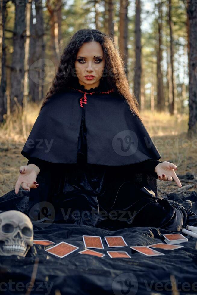 Witch in black dress with cape and hood. Posing in pine forest. Sitting on dark blanket whith fortune-telling cards and skulls on it. Full length. photo