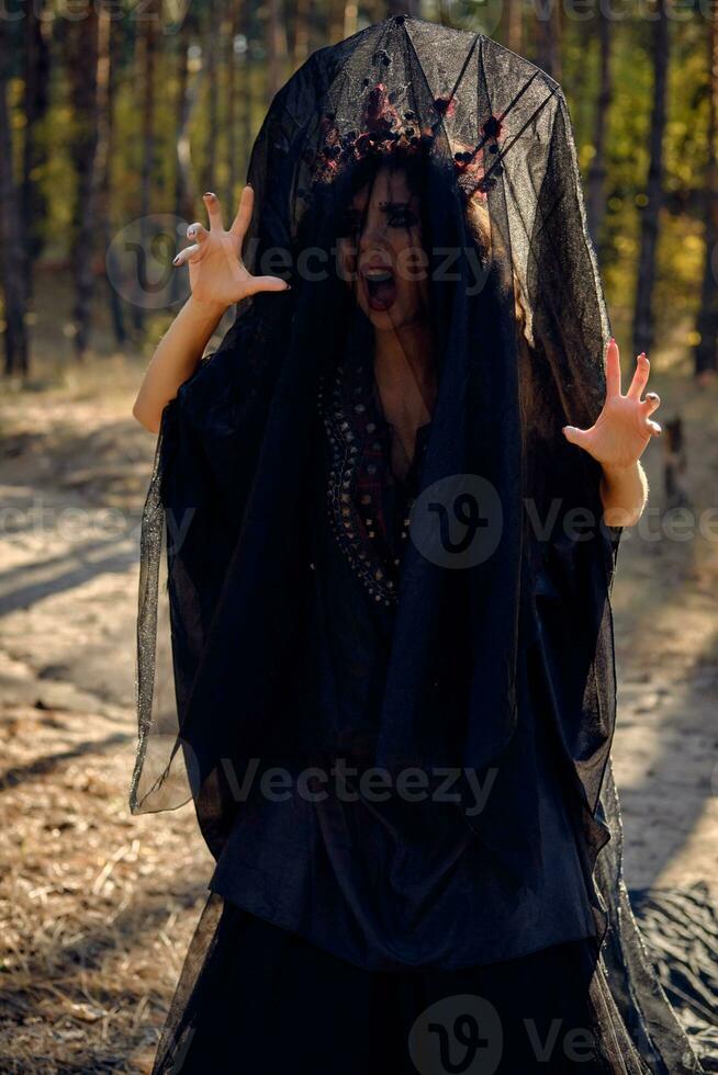 Witch in black, long dress, with red crown in her long, curly hair under a black veil. Posing in pine forest. Spells, magic and witchcraft. Close-up. photo
