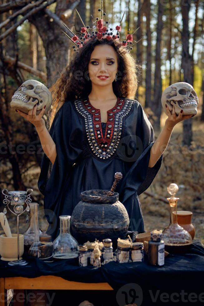 Witch in black, long dress, with red crown in her long, curly hair. Posing in pine forest. Holding skulls. Spells, magic and witchcraft. Close-up. photo