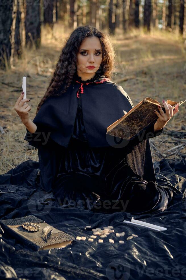 Witch in black dress with cape and hood. Posing in pine forest. Sitting on dark blanket whith books, candles and runes on it. Full length. photo