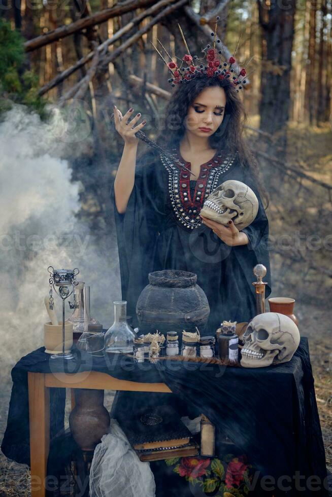 Witch in black, long dress, with red crown in her hair. Posing in pine forest, holding skull, making potion. Spells and witchcraft. Close-up, smoke. photo
