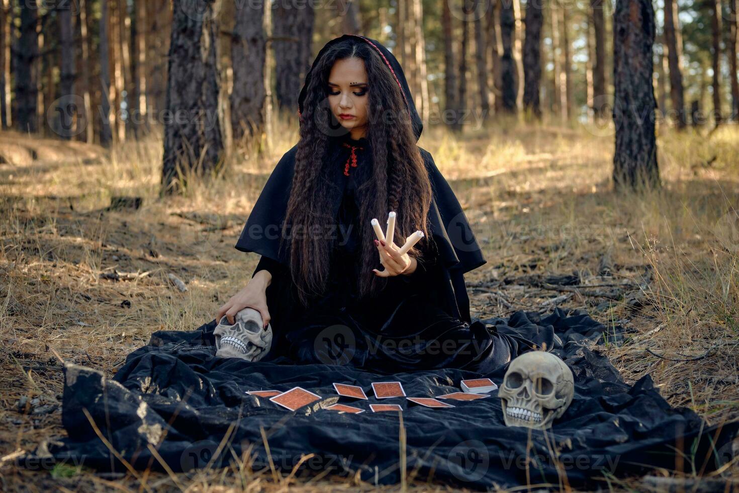 Witch in black dress with cape and hood. Posing in pine forest. Sitting on dark blanket whith fortune-telling cards and skulls on it. Full length. photo