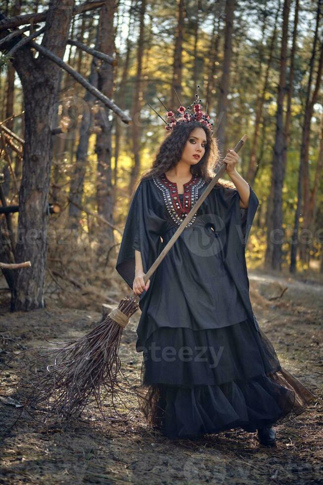 Witch in black, long dress, with red crown in her long hair. Posing with broom in pine forest. Spells, magic and witchcraft. Full length portrait. photo