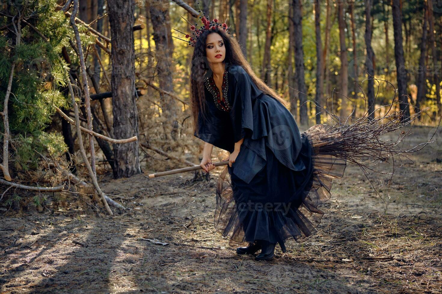 Witch in black, long dress, with red crown in her long hair. Posing sitting on broom in pine forest. Spells, magic and witchcraft. Full length. photo