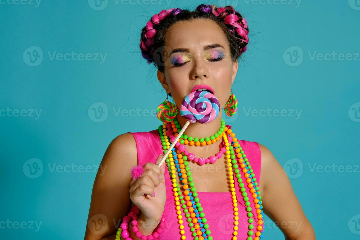 Lovely girl with a multi-colored braids hairstyle and bright make-up, posing in studio against a blue background, holding a lollipop in her hand. photo