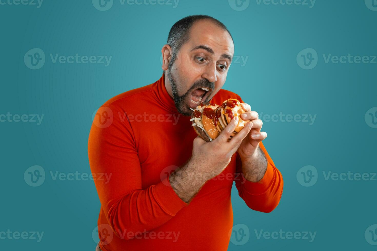 Close-up portrait of a middle-aged man with beard, dressed in a red turtleneck, posing with burgers against a blue background. Fast food. photo