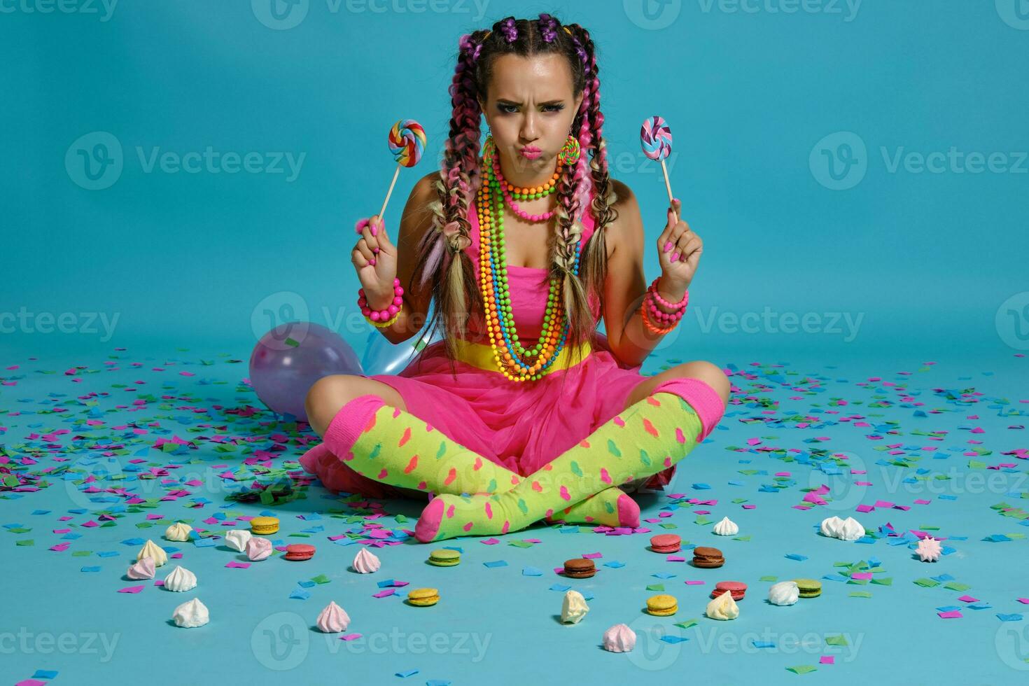 encantador niña con un multicolor trenzas peinado y brillante constituir, posando en estudio con chupete, aire globos y papel picado en contra un azul antecedentes. foto