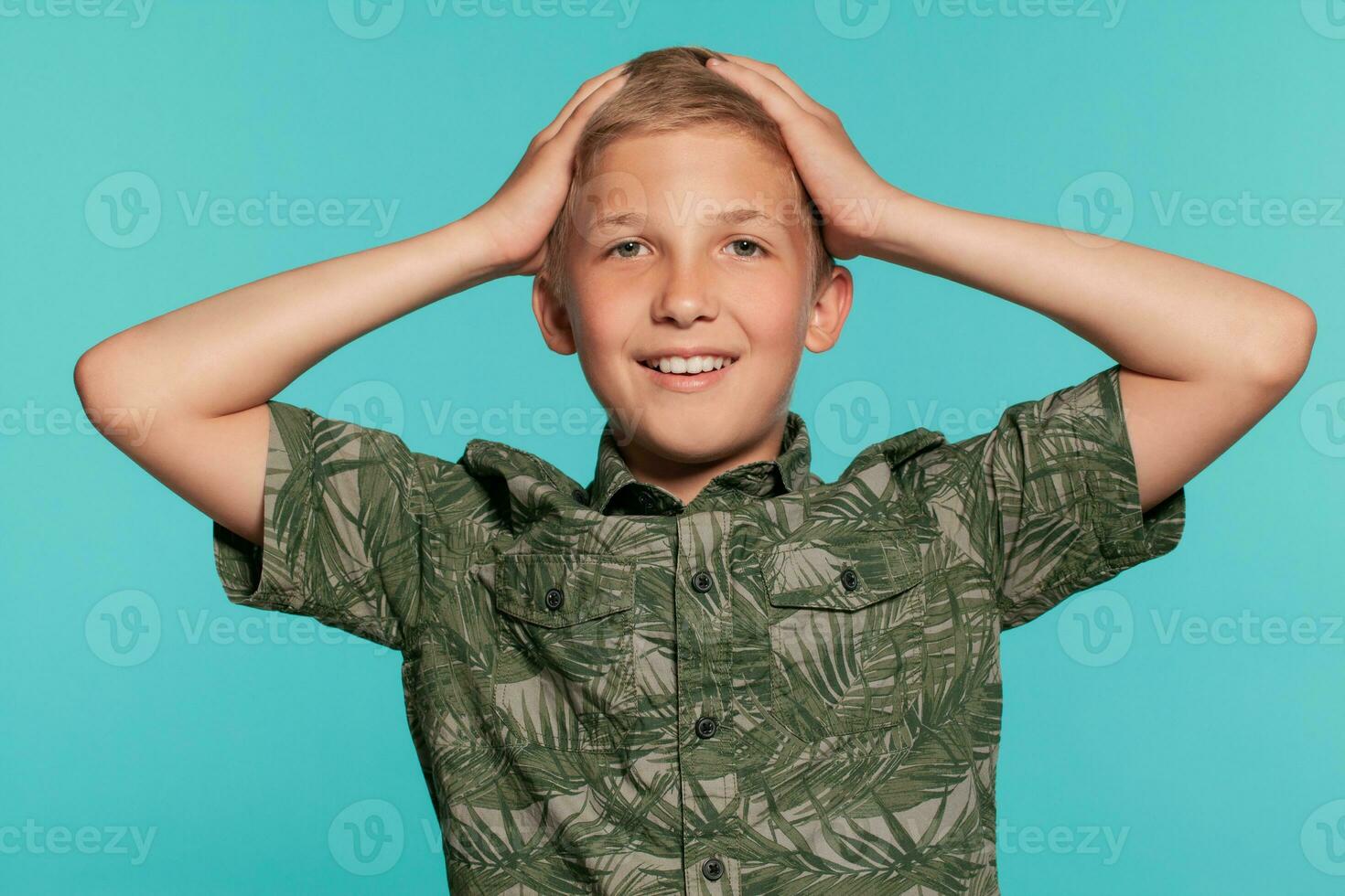 Close-up portrait of a blonde teenage boy in a green shirt with palm print posing against a blue studio background. Concept of sincere emotions. photo