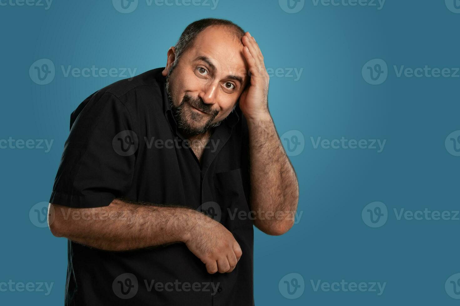 de cerca retrato de un moreno de edad mediana hombre con barba, vestido en un negro camiseta y posando en contra un azul antecedentes. foto