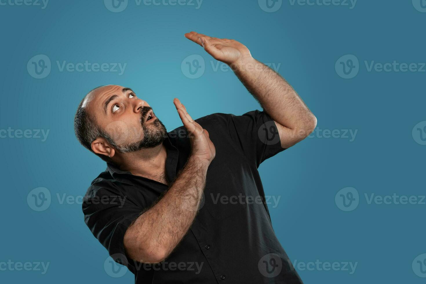 Close-up portrait of a brunet middle-aged man with beard, dressed in a black t-shirt and posing against a blue background. photo