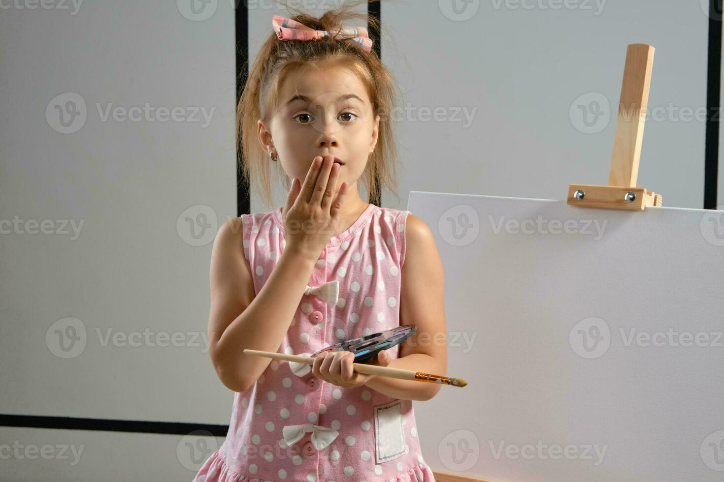 Little girl artist in a pink dress is standing behind easel and painting with brush on canvas at art studio with white walls. Medium close-up shot. photo