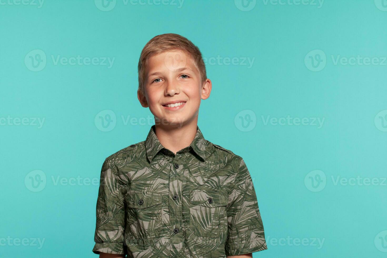 Close-up portrait of a blonde teenage boy in a green shirt with palm print posing against a blue studio background. Concept of sincere emotions. photo