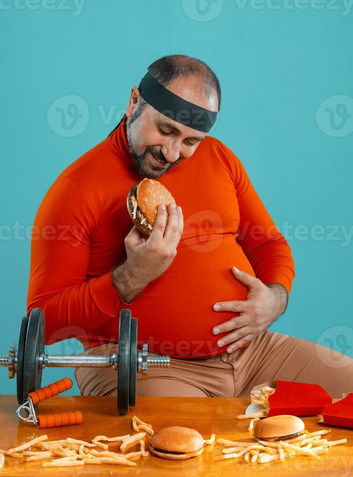 Middle-aged man with beard, dressed in a red turtleneck, headband, posing with burgers and french fries. Blue background. Close-up. Fast food. photo