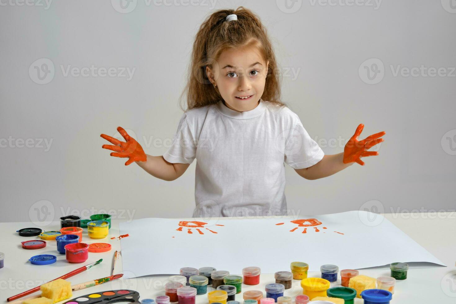 Little girl in white t-shirt sitting at table with whatman and colorful paints, showing her painted hands. Isolated on white. Medium close-up. photo