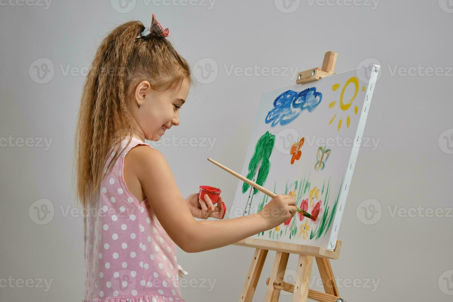 Little girl artist in a pink dress is standing behind easel and painting with brush on canvas, isolated on white studio background. Medium close-up. photo