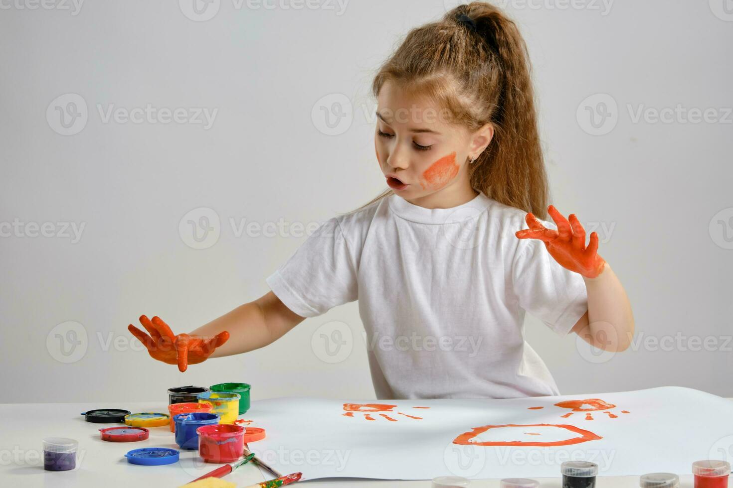 Little girl in white t-shirt sitting at table with whatman and colorful paints, painting on it with her hands. Isolated on white. Medium close-up. photo