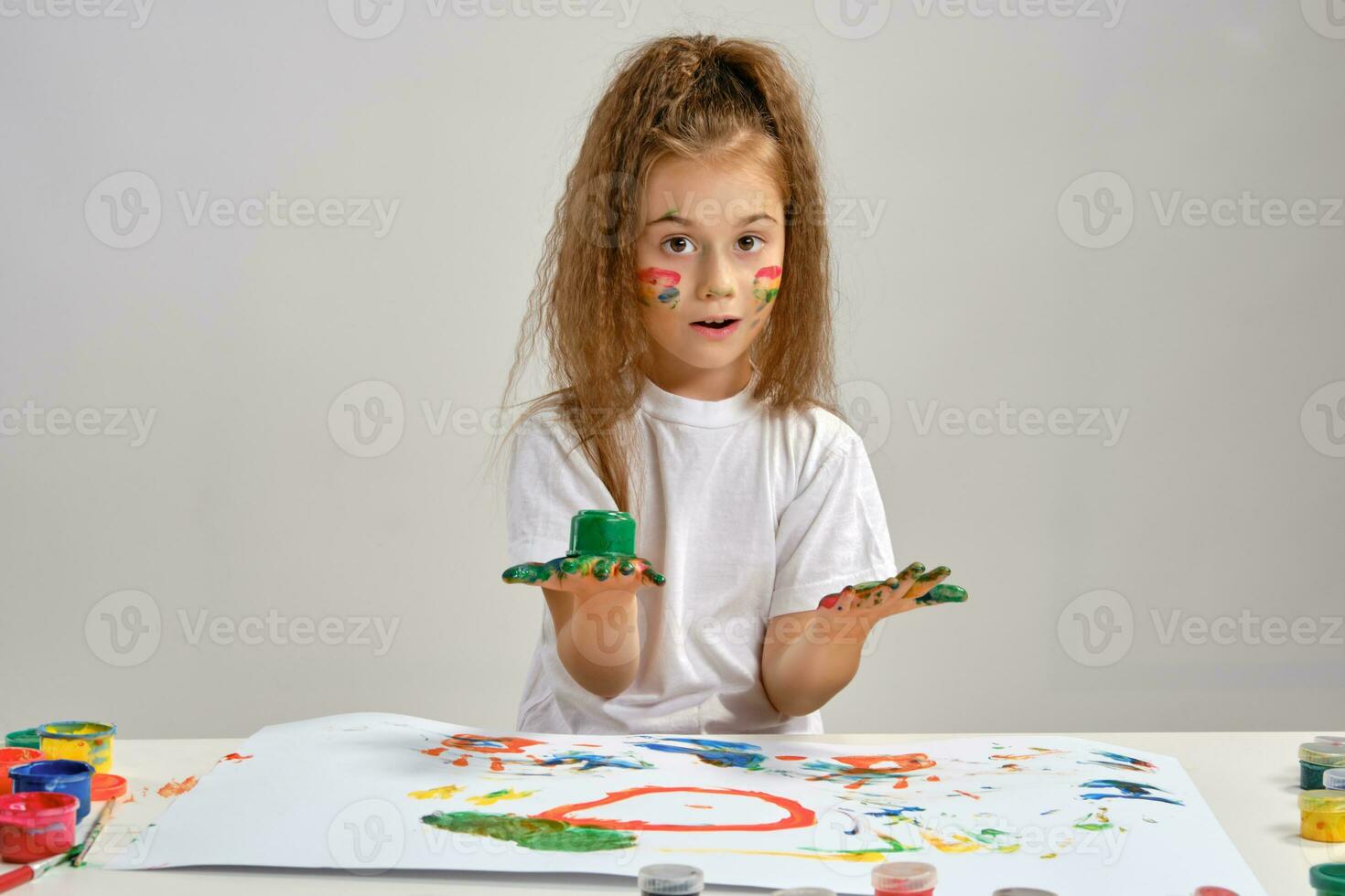 Little girl in white t-shirt sitting at table with whatman and paints on it, posing with painted face and hands. Isolated on white. Medium close-up. photo