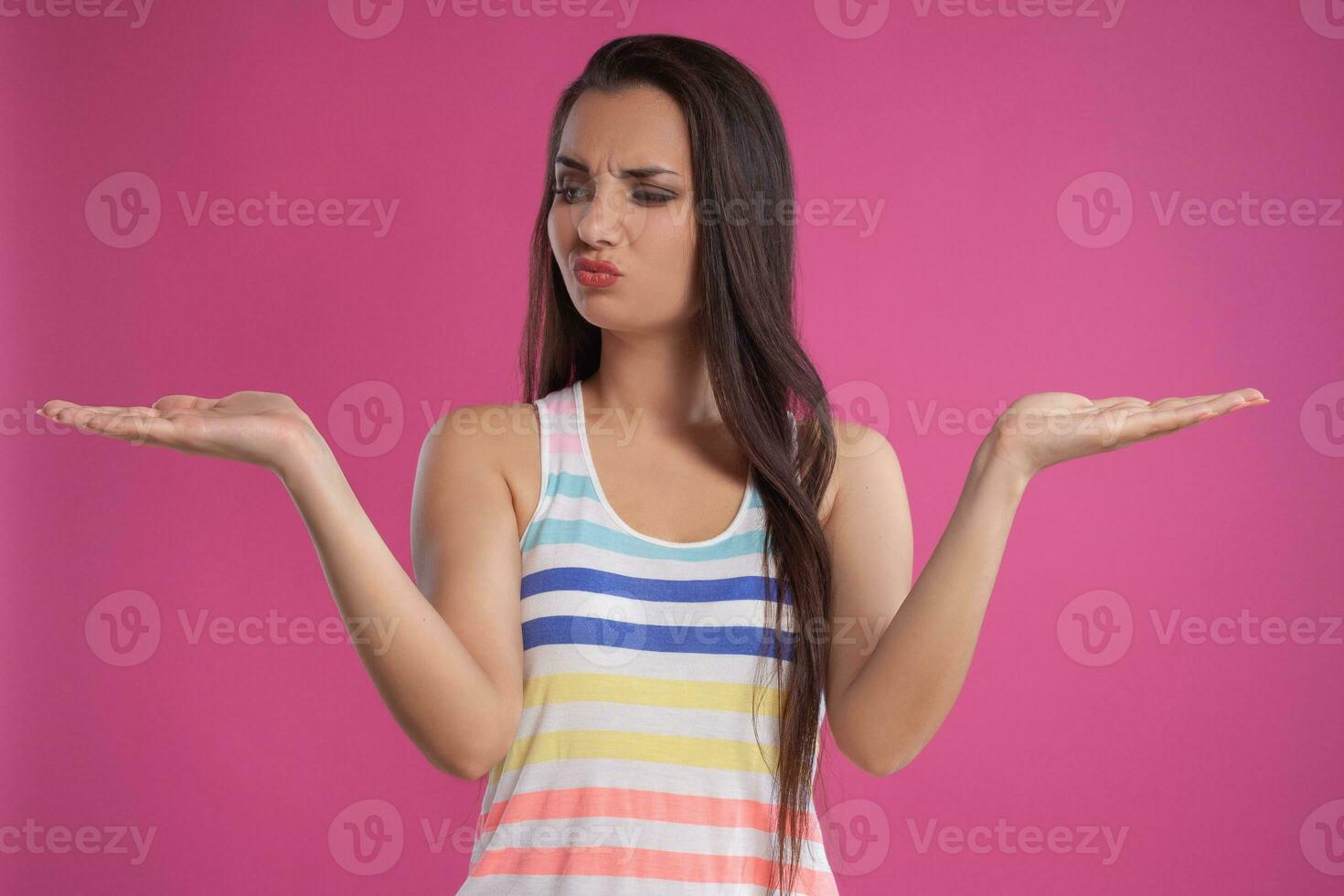 Brunette woman with long hair, dressed in colorful striped shirt, posing against pink studio background. Sincere emotions. Close-up. photo