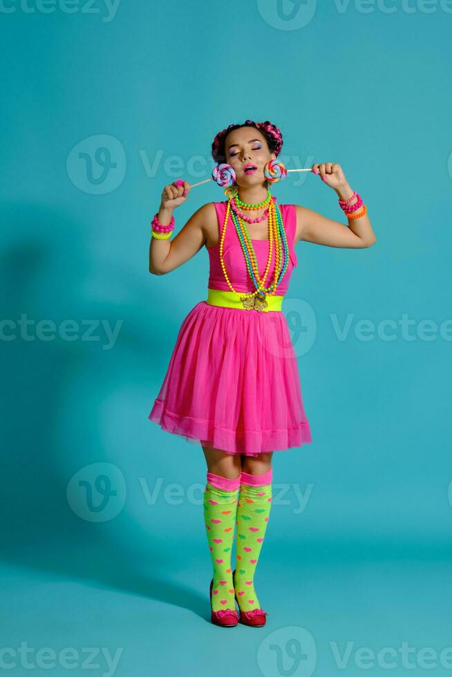 Lovely girl with a multi-colored braids hairstyle and bright make-up, posing in studio against a blue background, holding a lollipop in her hand. photo