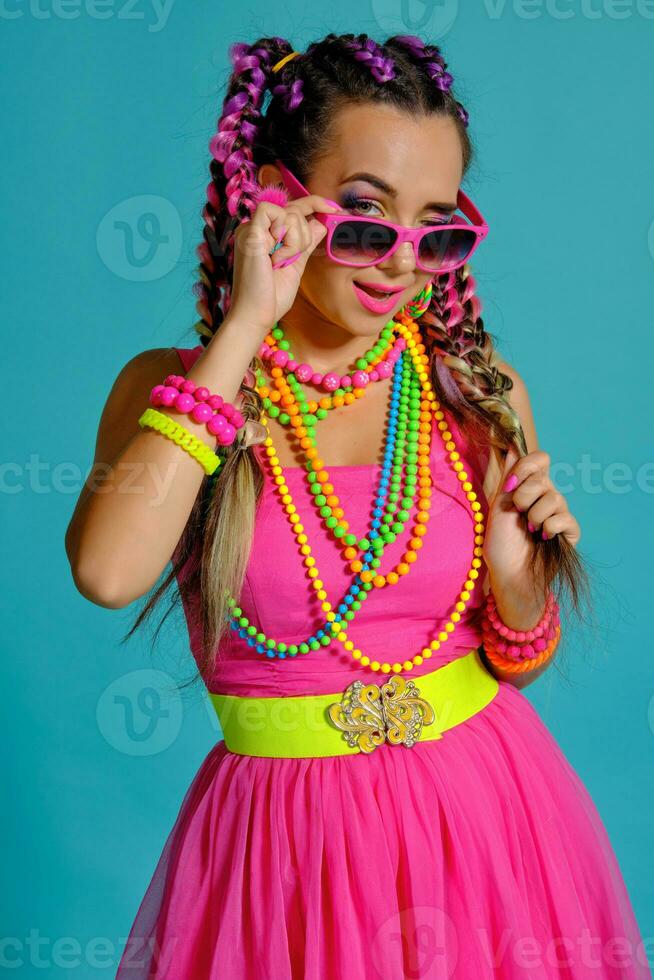 encantador niña con un multicolor trenzas peinado y brillante constituir, posando en estudio en contra un azul fondo, participación un pirulí en su mano. foto