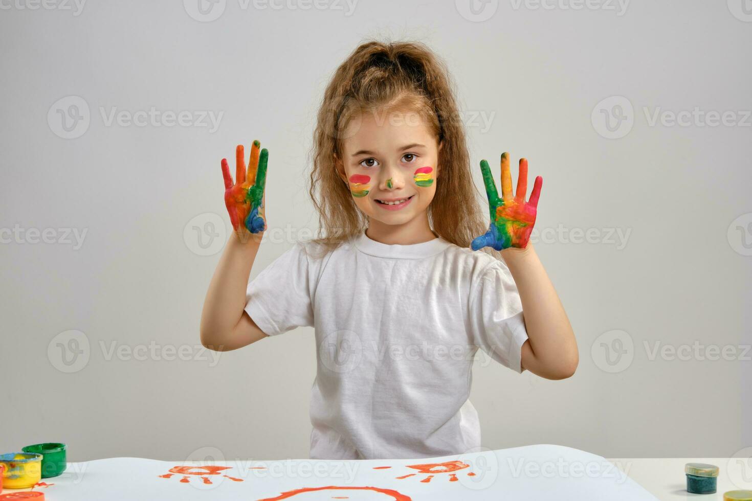 Little girl in white t-shirt standing at table with whatman and colorful paints, , showing painted hands, face. Isolated on white. Medium close-up. photo