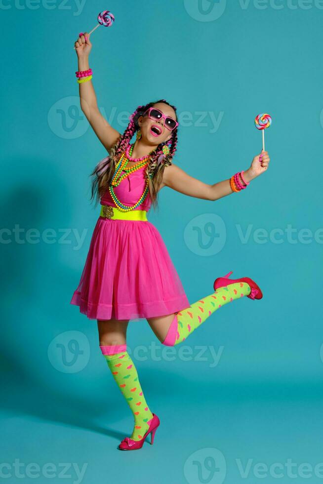 Lovely girl with a multi-colored braids hairstyle and bright make-up, posing in studio against a blue background, holding a lollipop in her hand. photo