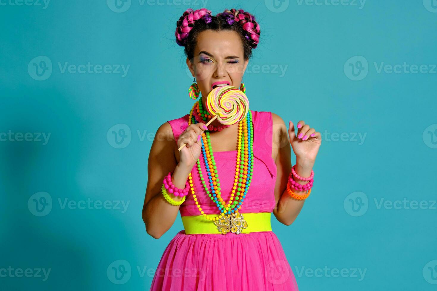 Lovely girl with a multi-colored braids hairstyle and bright make-up, posing in studio against a blue background, holding a lollipop in her hand. photo