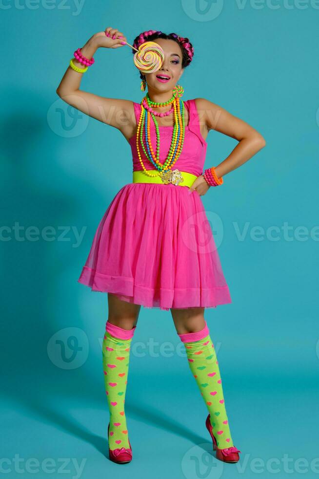 Lovely girl with a multi-colored braids hairstyle and bright make-up, posing in studio against a blue background, holding a lollipop in her hand. photo