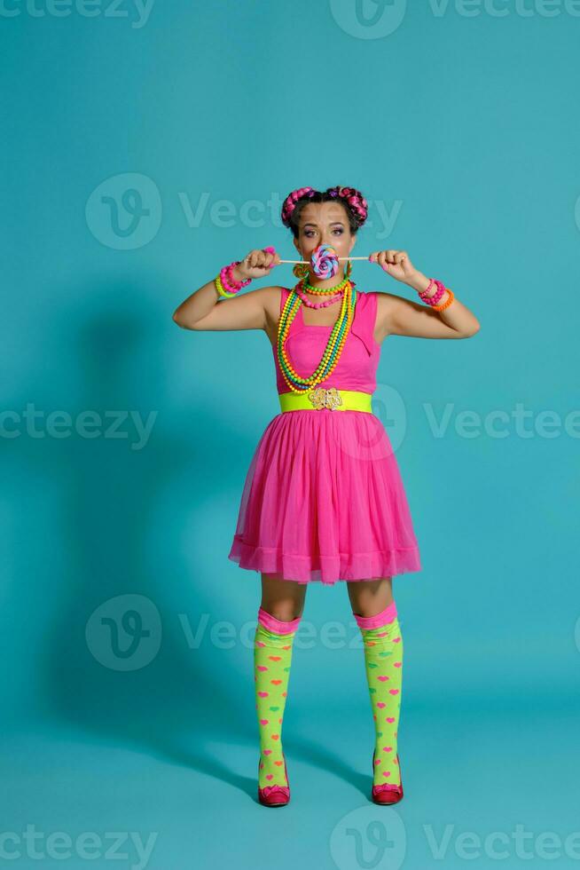 Lovely girl with a multi-colored braids hairstyle and bright make-up, posing in studio against a blue background, holding a lollipop in her hand. photo